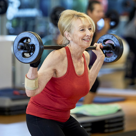 Photo of a woman wearing a red top in a group fitness studio smiling while lifting weights